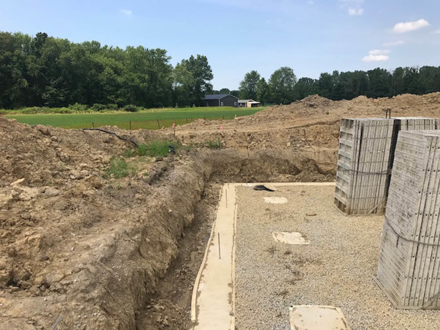 Excavated foundation surrounded by dirt and grass, concrete retaining wall, scattered stones, trees bordering the site, cloudy sky overhead