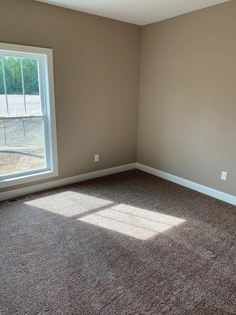 Carpeted bedroom with white walls, large window featuring metal bars, brown trim, and forest view outside