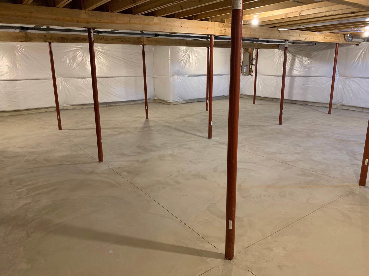 Open room with exposed wooden ceiling beam, white painted wall, concrete floor, metal support pole, recessed ceiling light, and white plastic sheeting covering part of the wall