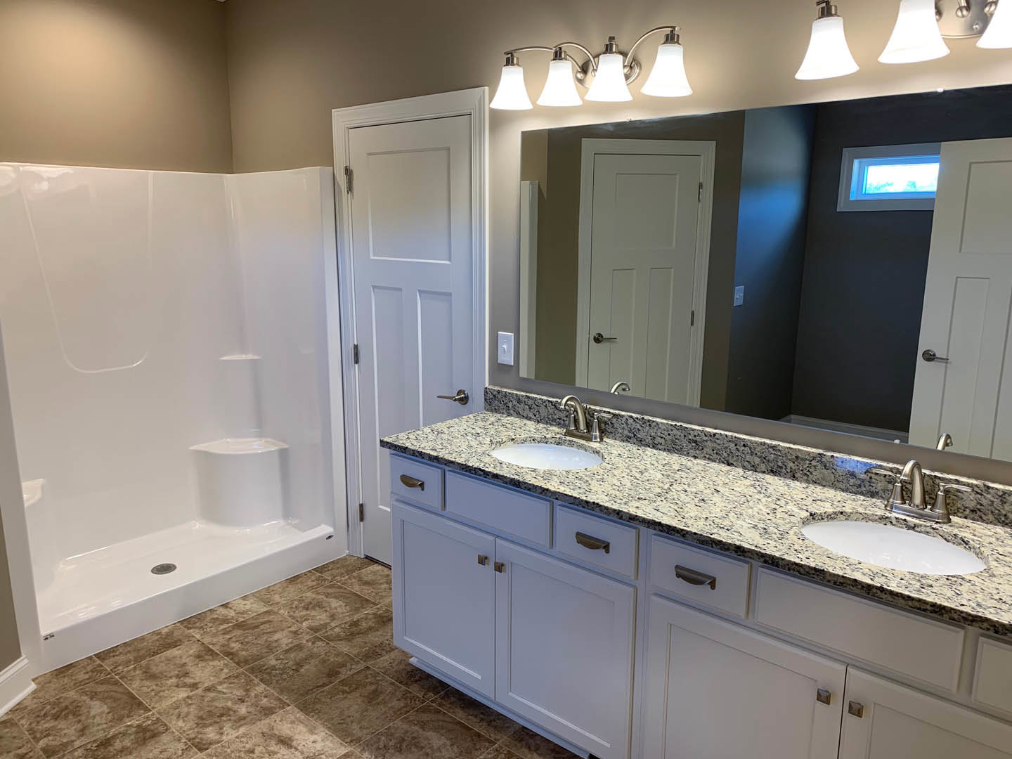 White tile shower with chrome drain, adjacent white vanity with undermount sink and silver faucet, white paneled door with silver doorknob, window with trim letting in natural