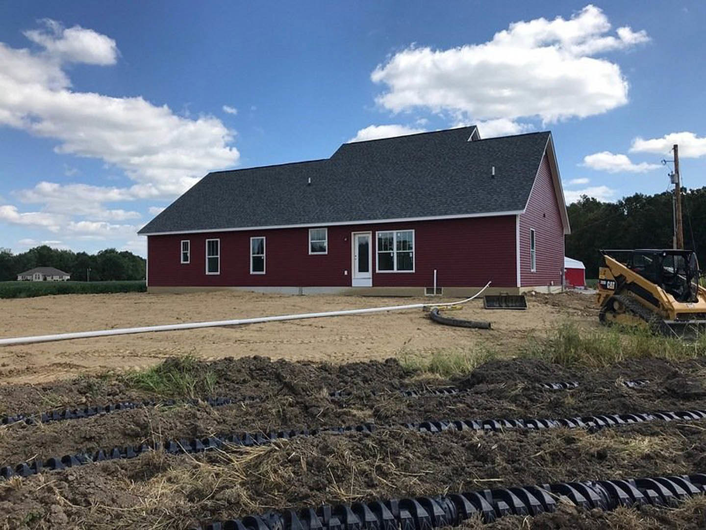 Red house with white door and gabled roof, yellow tractor parked on dirt construction site, grassy lot, blue sky with scattered clouds.
