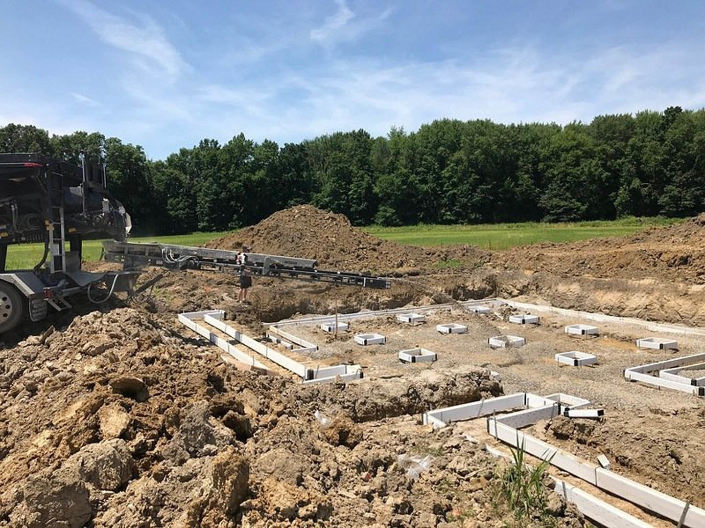 Excavator parked on dirt foundation at residential construction site, surrounded by soil, grass, and trees under a partly cloudy blue sky