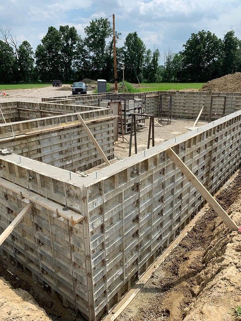 Concrete foundation under construction with exposed rebar, surrounding soil, and a partially built brick wall; telephone pole and power lines visible in background, trees and