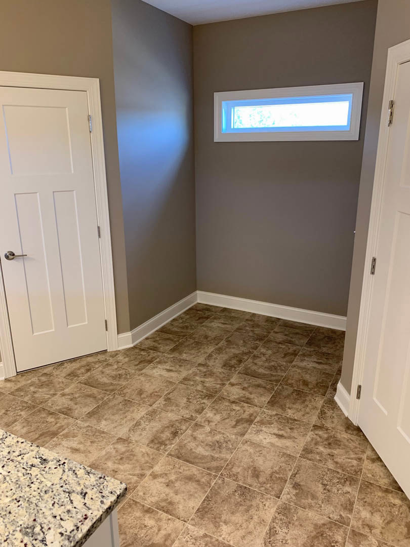 Brown tile flooring with white baseboard trim, two white paneled doors with metal hinges, a window with white casing, smooth plaster walls, and a light ceiling in a residential