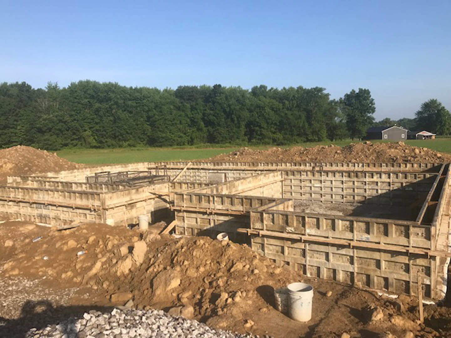 Partially built residential structure with exposed framing, dirt construction site, scattered white bucket, and dense trees under clear blue sky in background