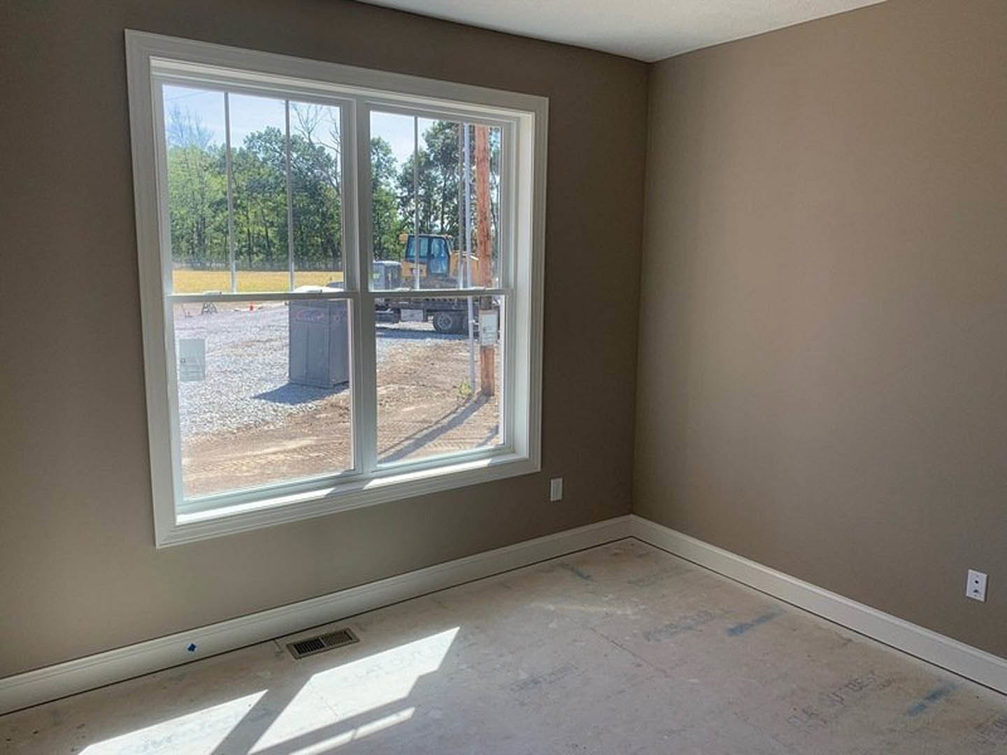 Concrete floor with a rectangular floor vent, white plaster walls, large window overlooking construction site, natural daylight illuminating the space