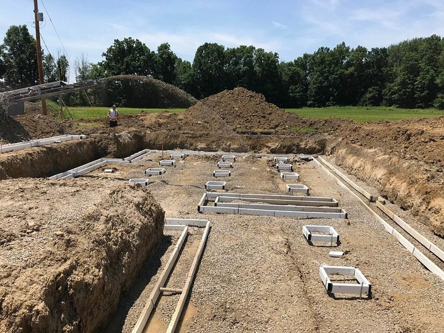 Construction site with exposed soil, large dirt mound, trench, scattered rocks, and trees in background; man in white shirt standing near foundation area under cloudy sky