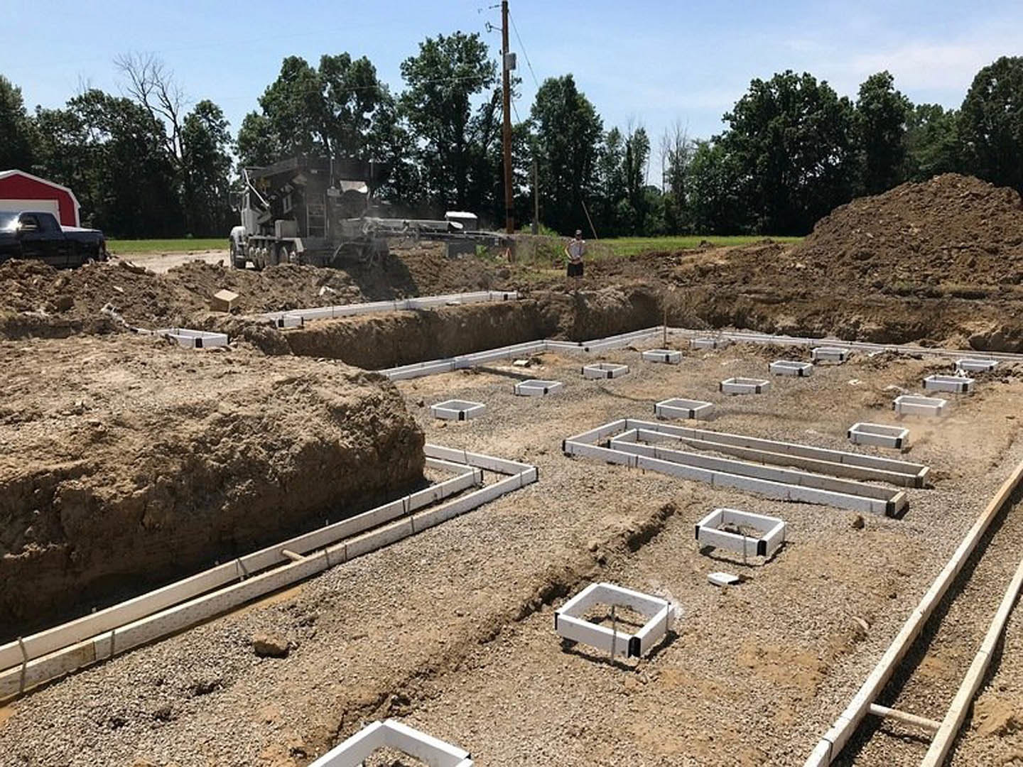 Bulldozer and black truck parked on dirt construction site beside partially built foundation with white square frames, exposed pipes, and telephone pole; trees and blue sky in