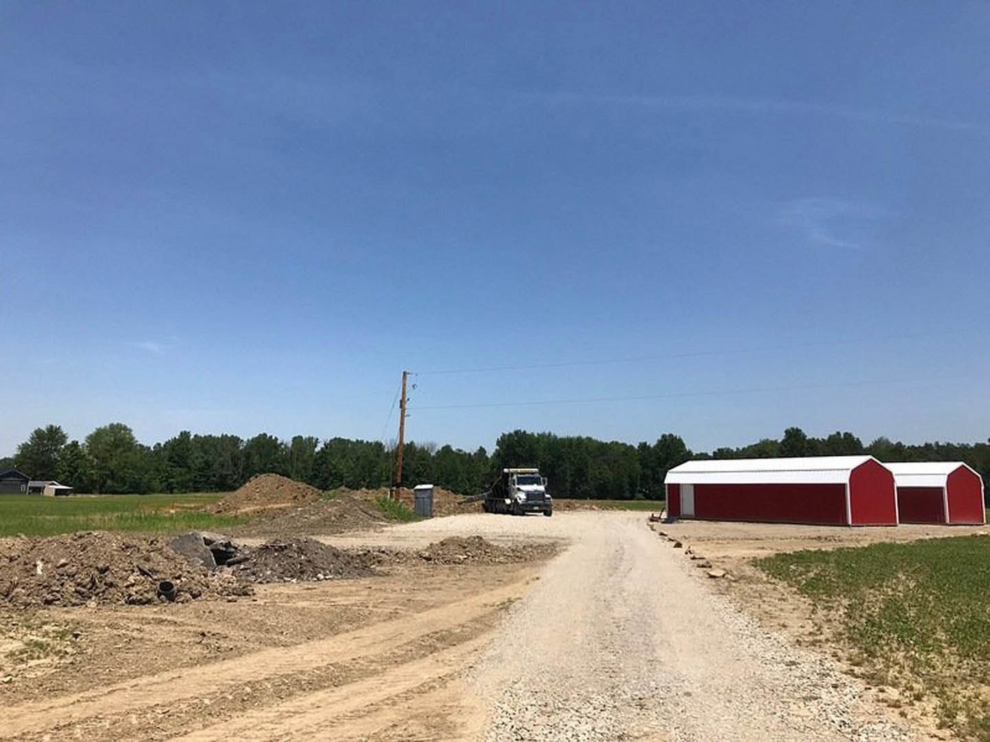Dirt road lined with grass and dirt, red and white shed, parked truck, blue sky with scattered clouds, rural landscape
