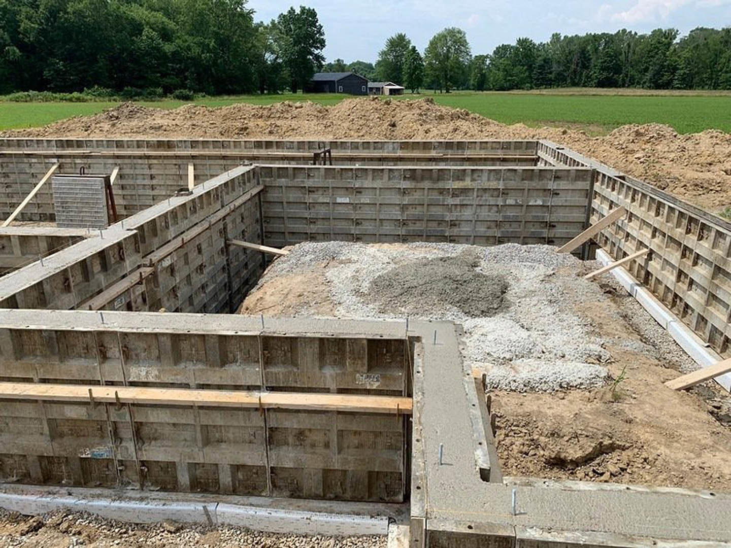 Concrete foundation under construction with exposed concrete walls, wooden beam, metal utility box, and gravel pile; wooded area and neighboring houses in background.