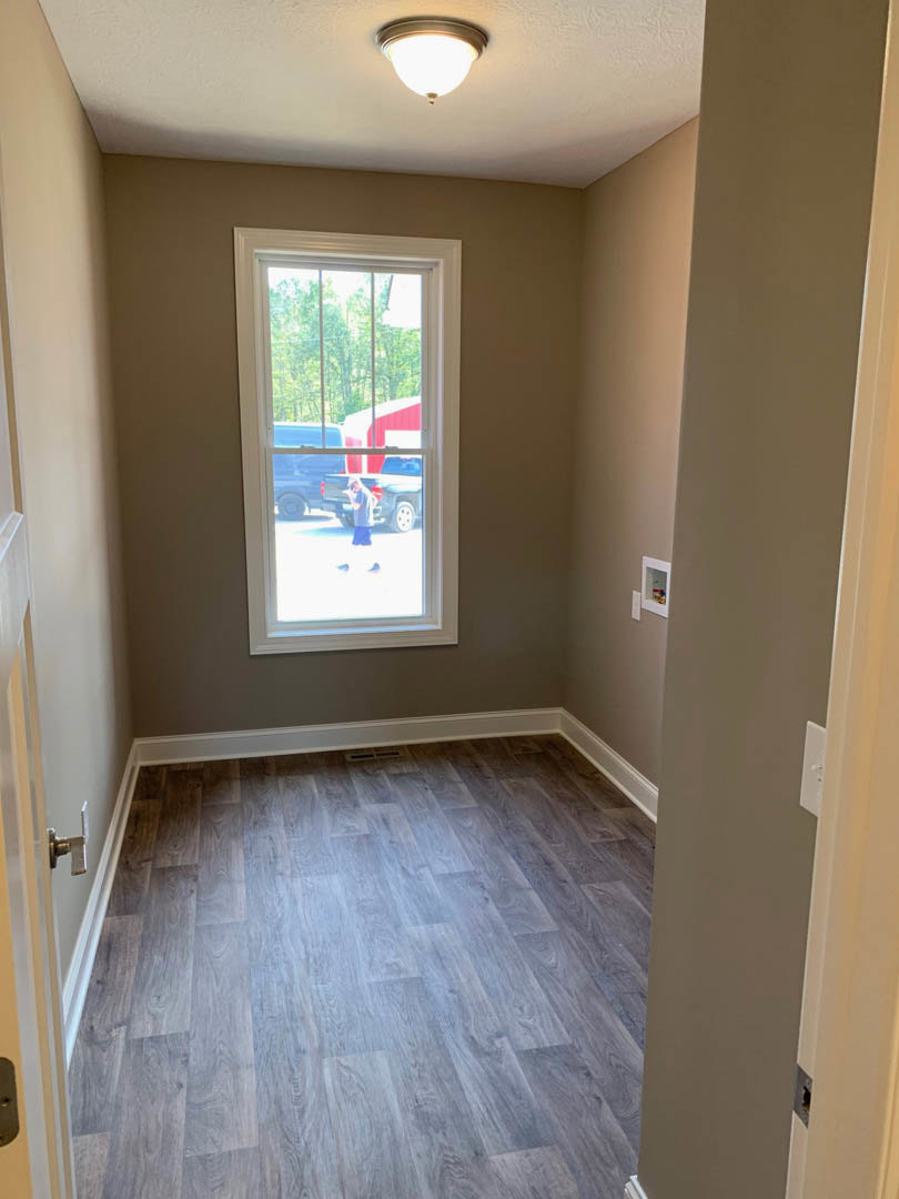 Sunlit room featuring wide plank wood flooring, large window with views of a red building and parked car, white walls, and modern ceiling light fixture
