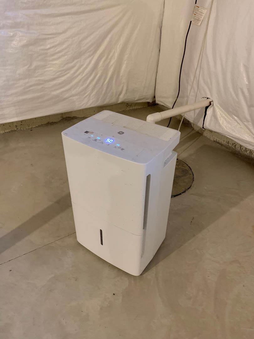 White rectangular home appliance with blue indicator lights mounted on a wall above a white pipe, set against a neutral interior background.