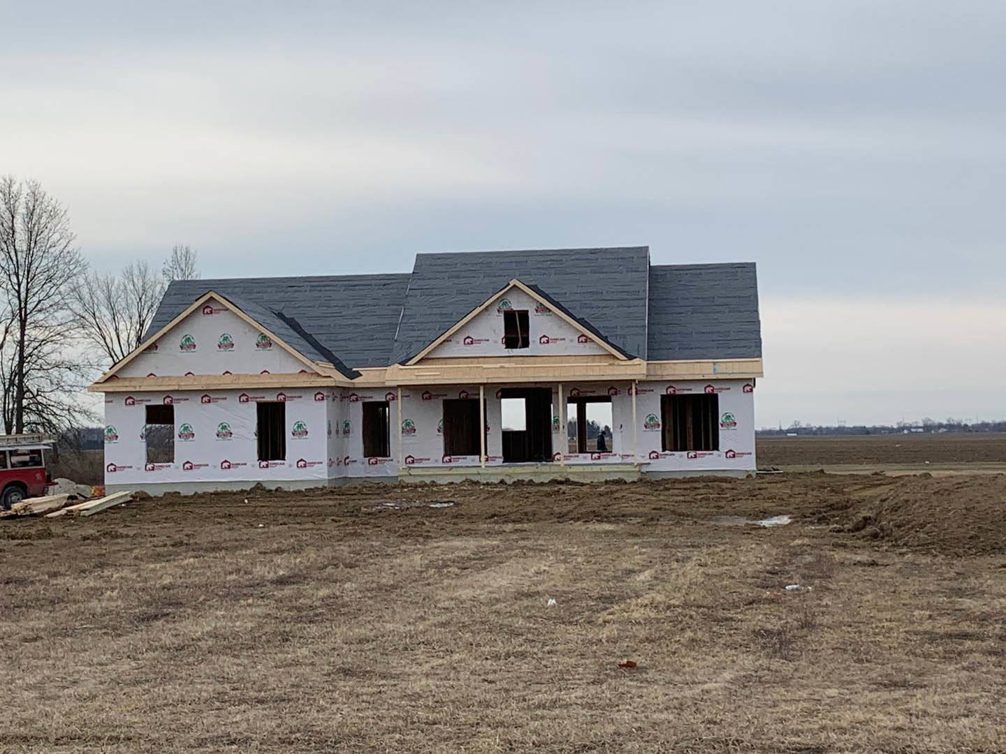 Partially built house with grey roof and exposed framing, surrounded by brown grass field, leafless tree nearby, red vehicle parked beside structure, cloudy sky overhead