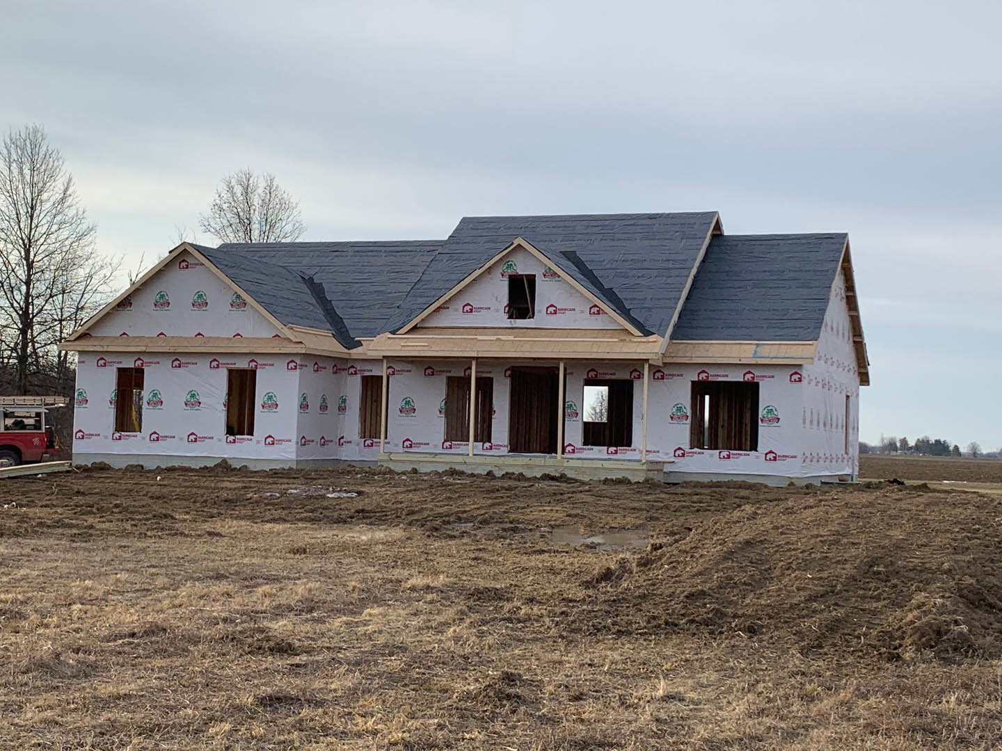 Partially built house with black roof and white siding, surrounded by dirt field, leafless tree nearby, red truck with ladder parked in front, Robert Frost Farm visible in