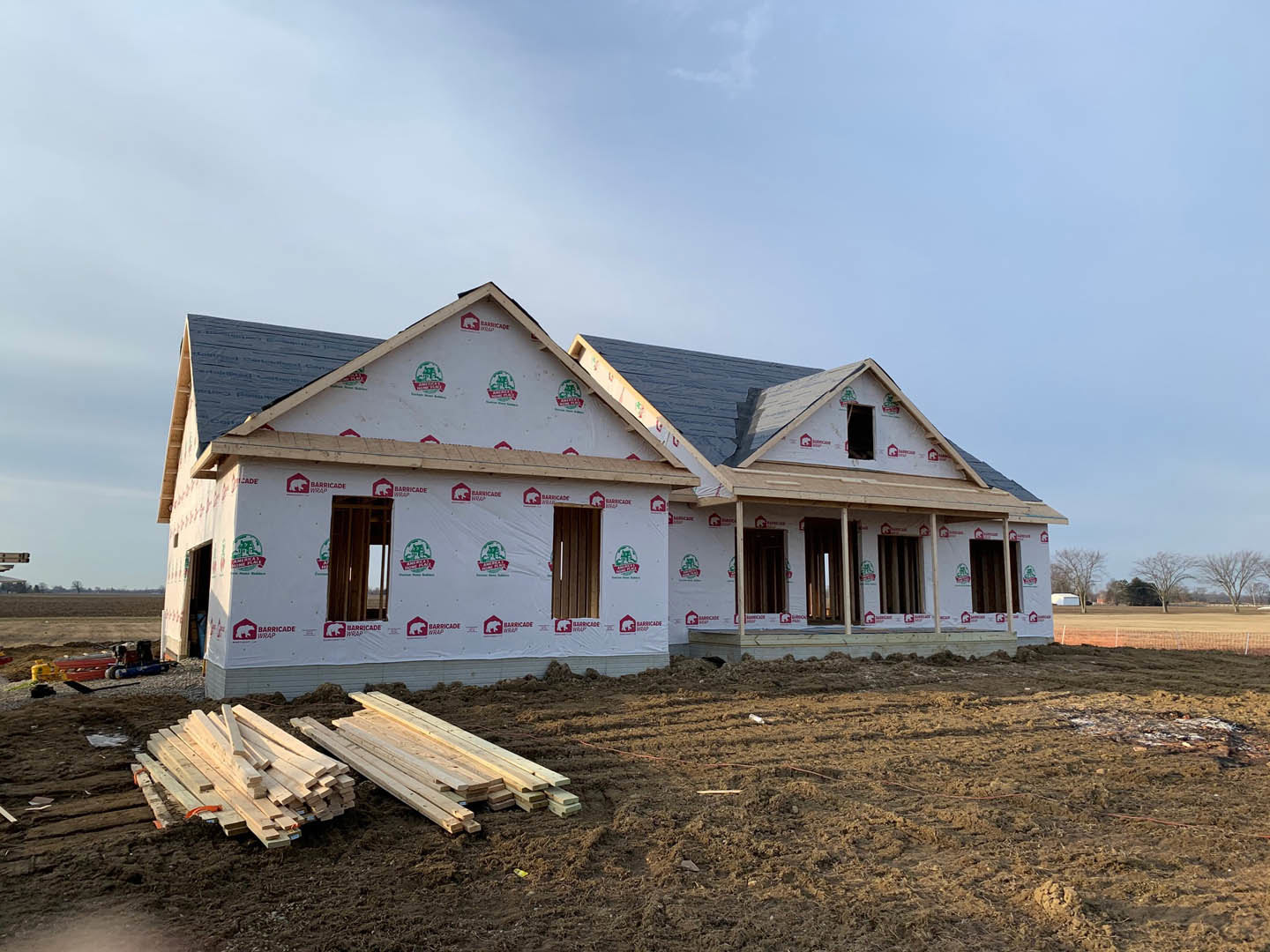 Framed house under construction with exposed wood planks, white protective covering, unfinished door opening, and stacked lumber on dirt ground