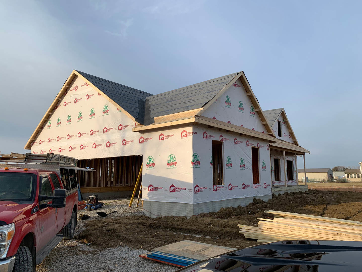 Partially built house with exposed framing, construction materials scattered on dirt ground, red pickup truck parked in front, ladder leaning against exterior wall, temporary sign