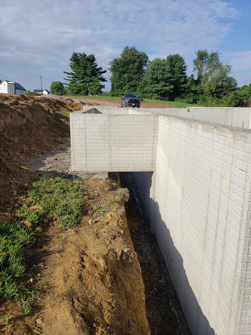 Concrete retaining wall with a circular hole in the ground, patchy grass and dirt along the base, black truck and car on nearby road, white brick wall section, group of trees under