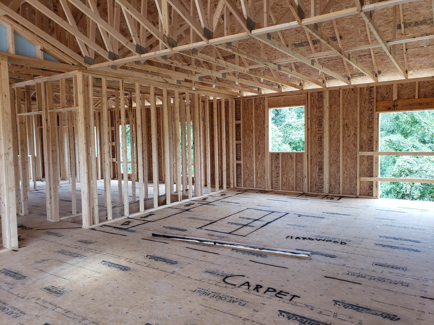 Open room with exposed wood ceiling beams, large window overlooking trees, unfinished floor with construction materials visible