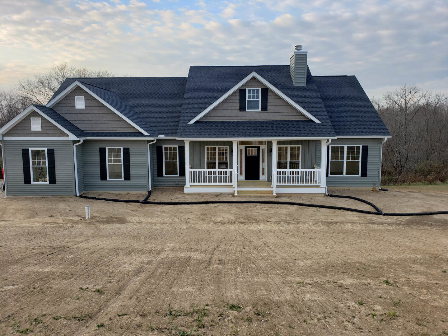 White two-story house with black roof, black front door with white trim, covered porch, large dirt yard with garden hose, white-framed windows, cloudy sky overhead
