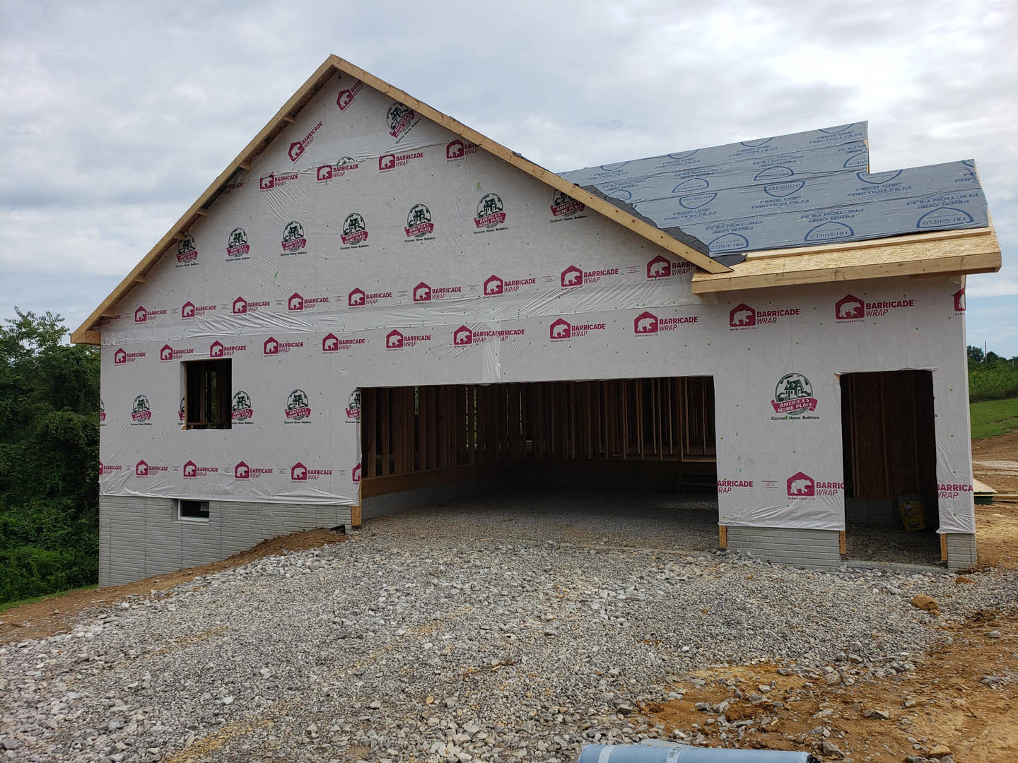 Framed custom home with attached garage, exposed wood beams, unfinished exterior walls, gravel pile in foreground, cloudy sky overhead, trees in background