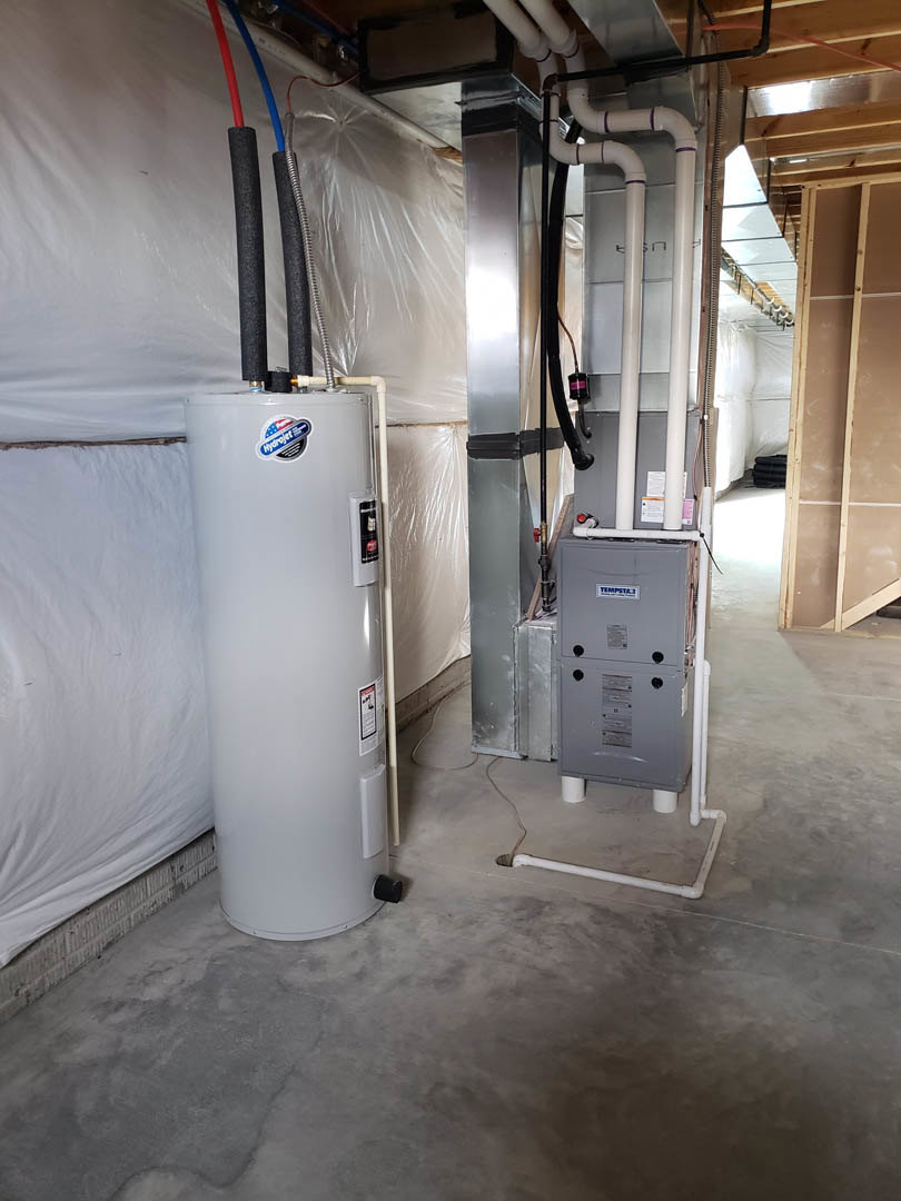 White cylindrical gas heater with labeled control panel, positioned against a wood-framed wall near a grey box and exposed pipe, resting on a light-colored floor.