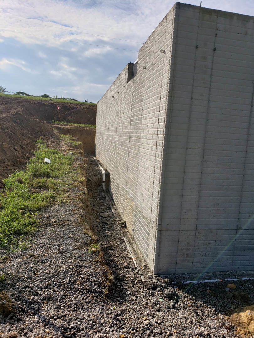 Red brick wall bordering a grassy ditch with exposed soil, cloudy sky overhead, and faint rainbow visible near the corner of the wall