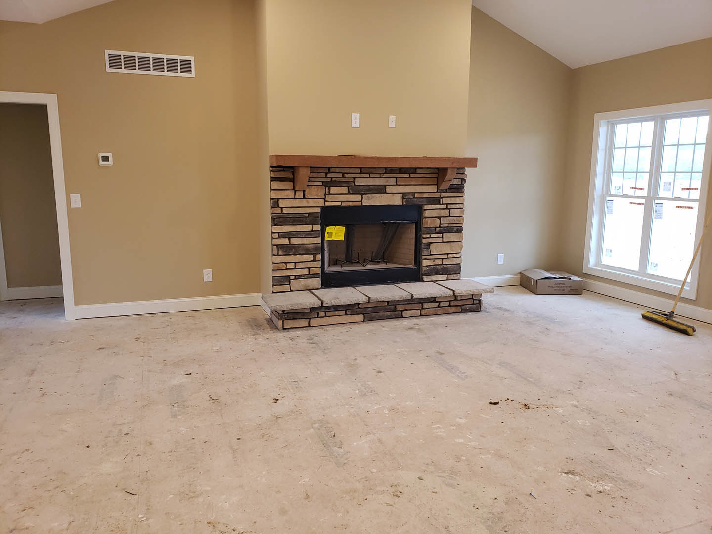 Stone fireplace with yellow sign on hearth, brown walls with white trim, large window with white casing, hardwood floor