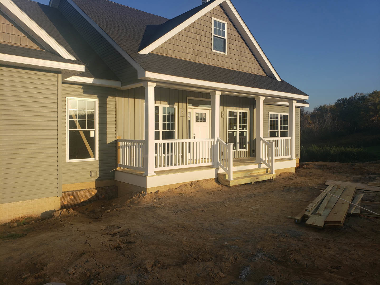 Partially built house with white porch railing, exposed wooden framing, white window frame, wood pallet and planks on dirt ground