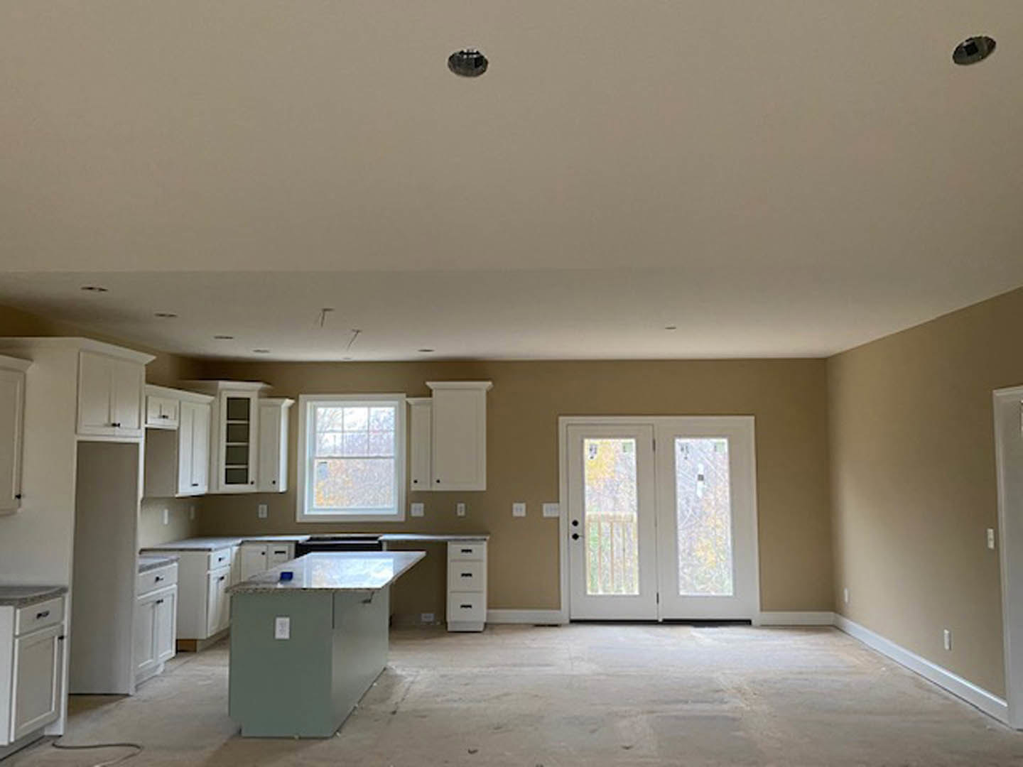 Open kitchen featuring a white countertop island, light wood cabinetry, glass-paneled double doors, white-framed window, pale flooring, and white walls with black hardware