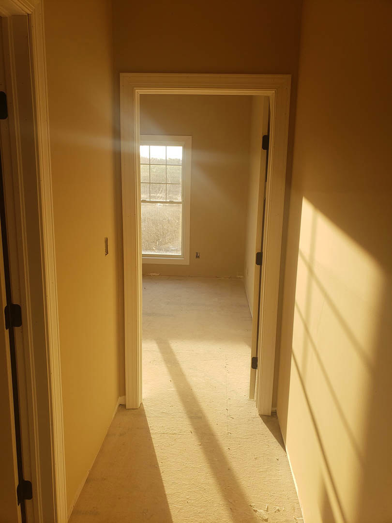 Hallway with light wood flooring, white plaster walls, open white door revealing adjacent room, sunlight streaming through window with white frame