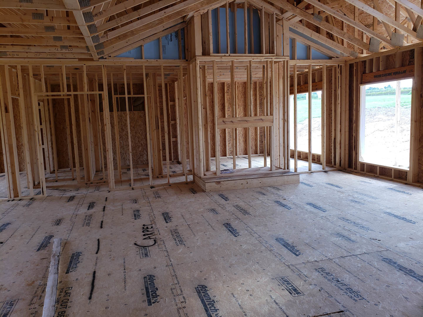 Wood-framed room with exposed beams, unfinished wood floor marked with black text, large window overlooking grassy yard