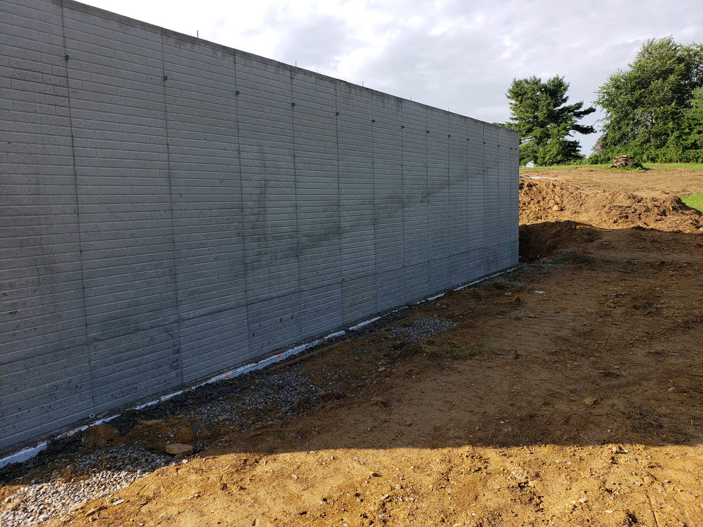 Grey brick exterior wall with circular holes, bordered by a dirt path and tall leafy trees under a partly cloudy sky.