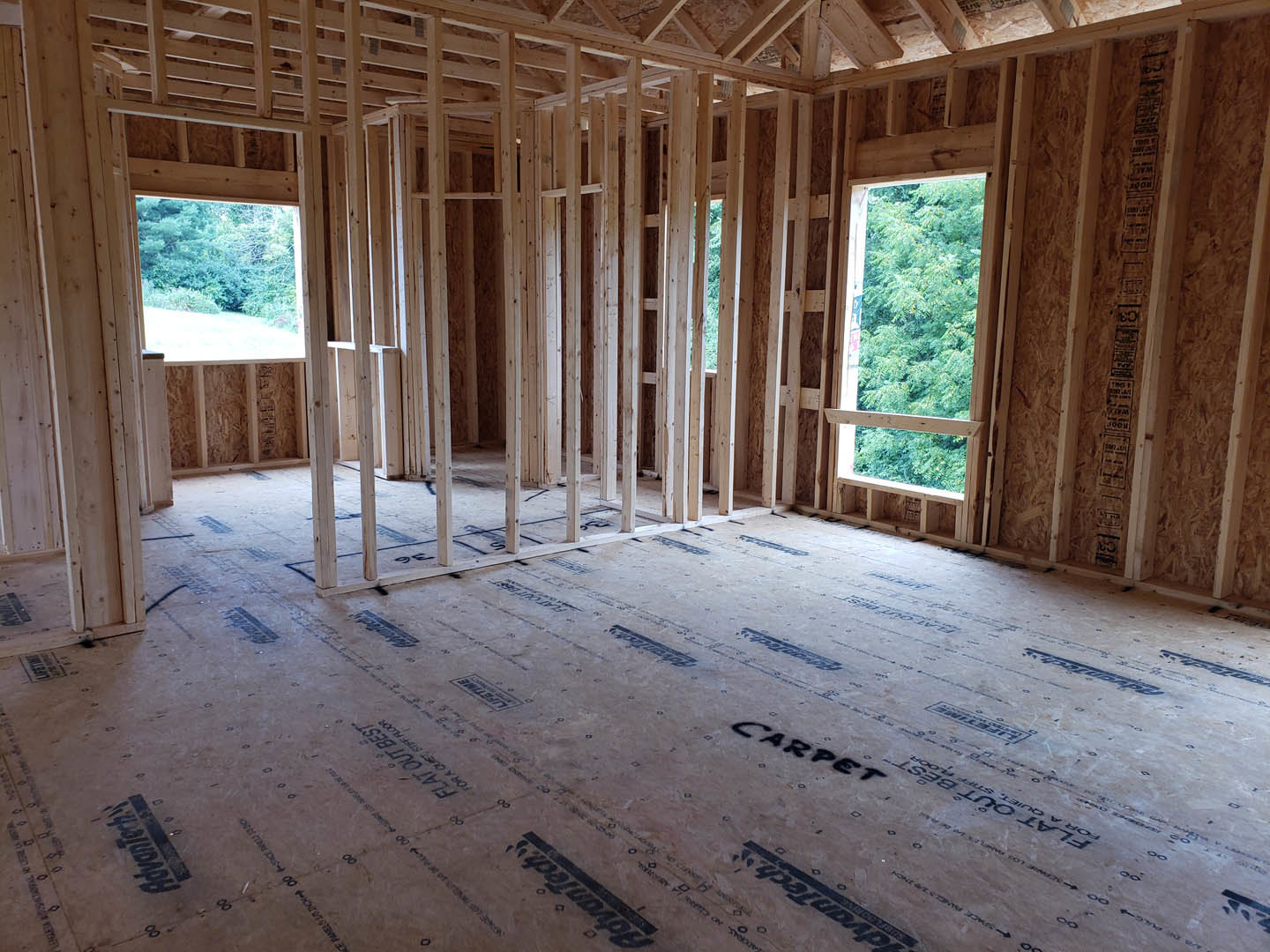 Wood-framed room under construction with exposed beams, unfinished wood floor, large window revealing trees outside