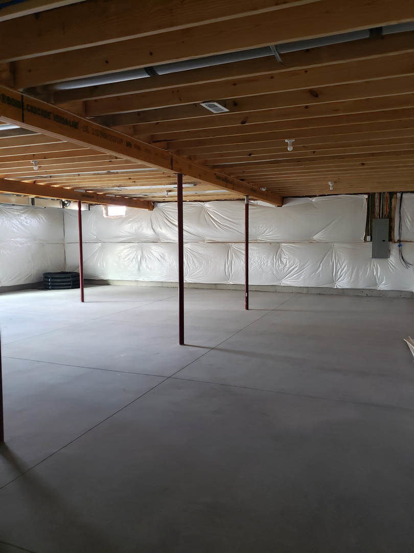 White wall with exposed wooden ceiling beams, concrete floor partially covered by a white sheet, black object mounted on wall, unfinished interior.