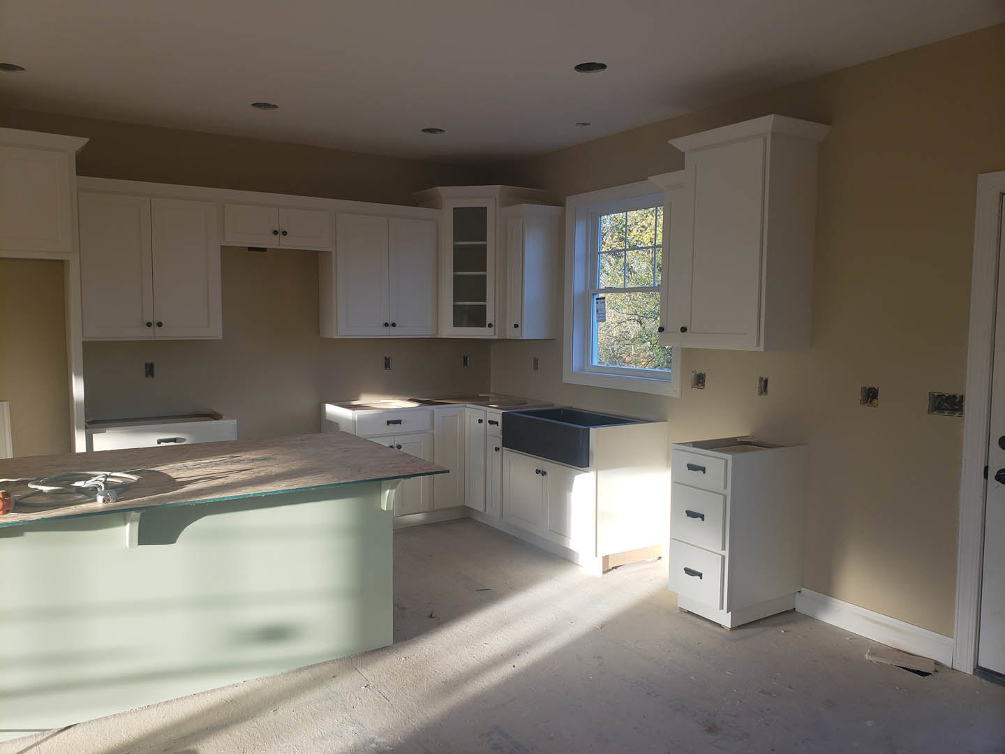 White kitchen with shaker cabinets, quartz countertops, stainless steel sink, glass-topped desk, black-framed open shelving, large window with signage, and light wood flooring.