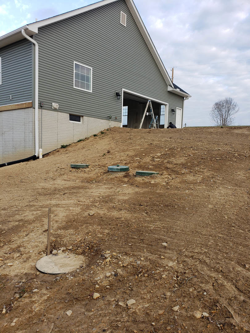 Two-story home with light siding, attached garage, multi-pane windows, leafless tree, dirt hill with round holes, circular object, ladder leaning against closed garage door, cloudy