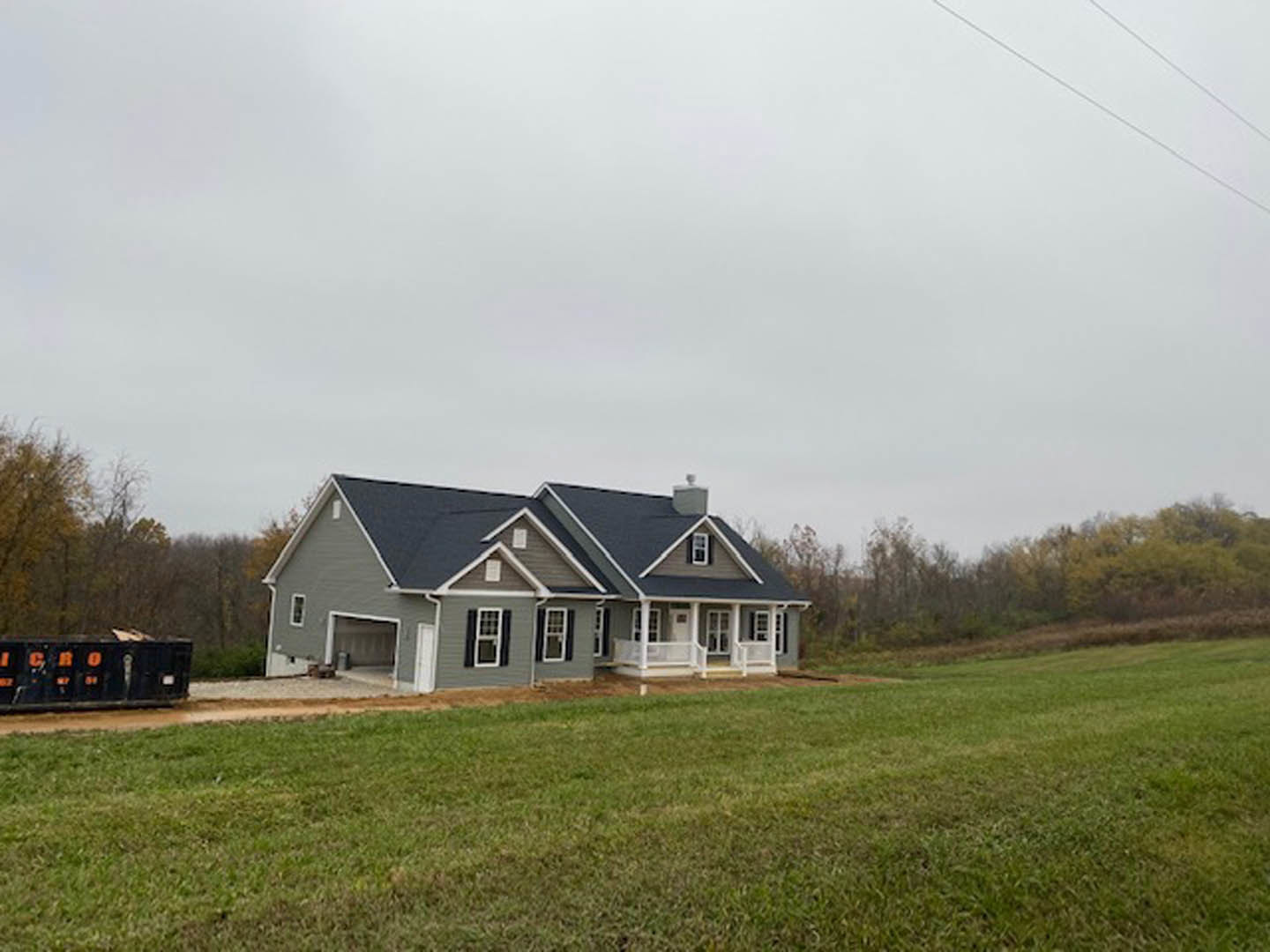 Two-story farmhouse with grey roof, grid windows, and expansive green lawn bordered by mature trees under a clear sky