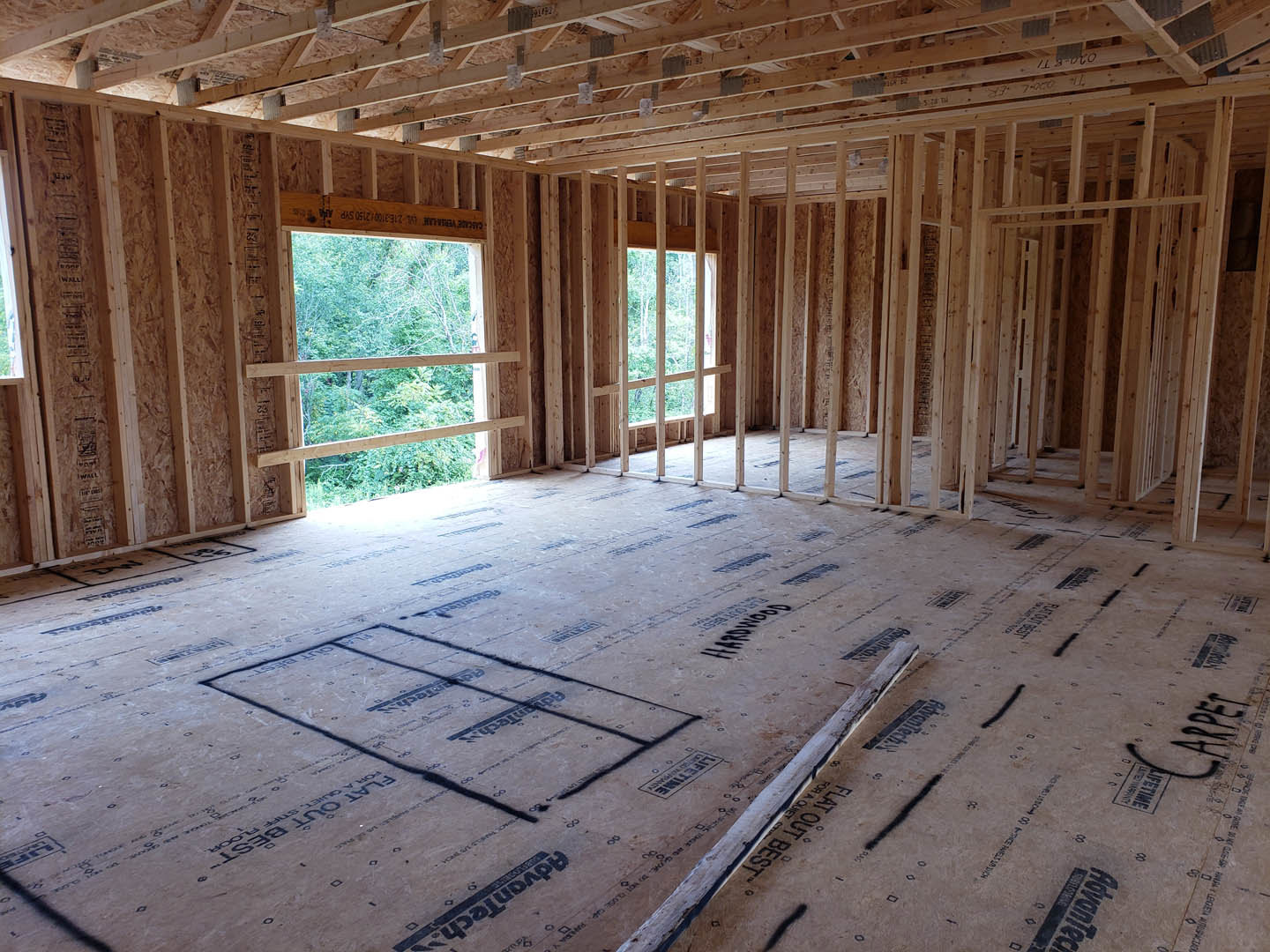 Living room with exposed wood ceiling beams, large windows framed in wood, carpeted floor, and construction materials visible.