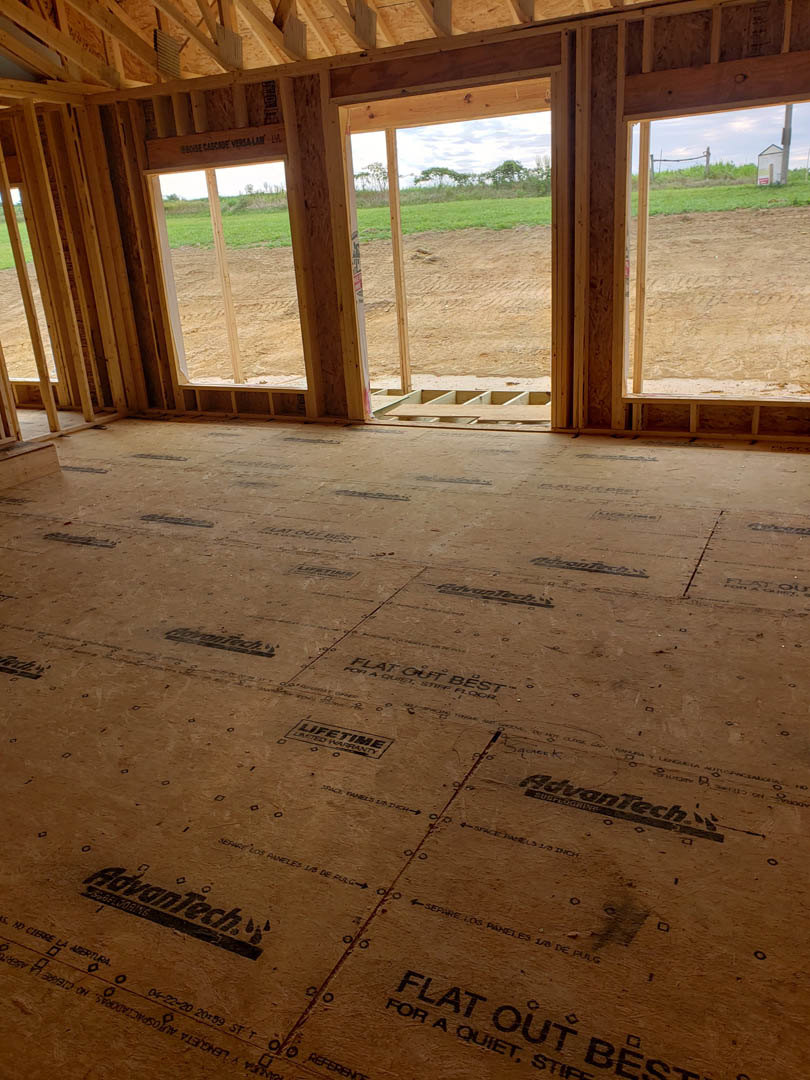 Sunlit room with unfinished plywood flooring, large window overlooking grassy field, handwritten logo sign visible in foreground