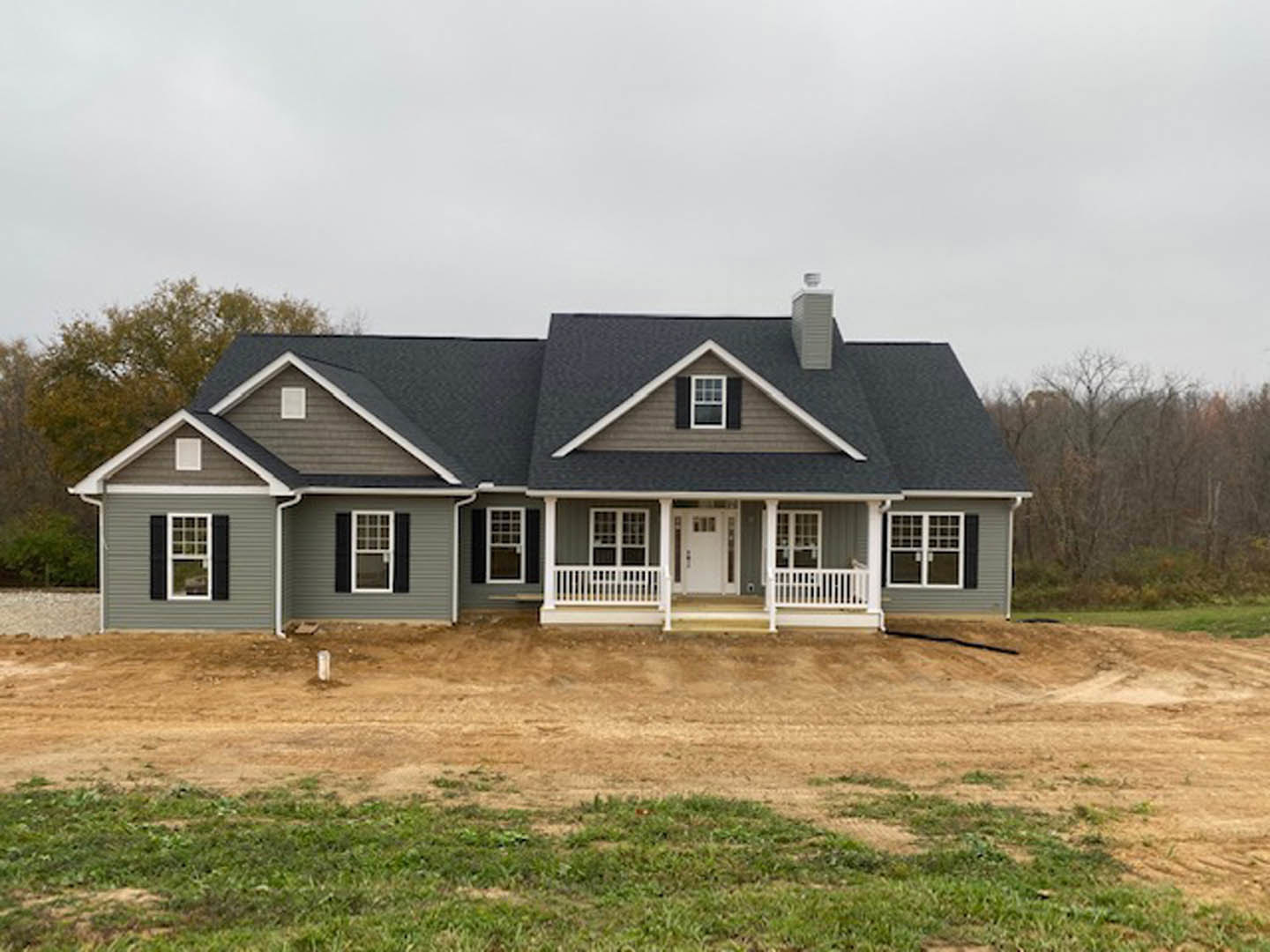 Partially built home surrounded by dirt and mature trees, featuring a white door with glass panels, white-framed windows, and white railings; cloudy sky overhead.