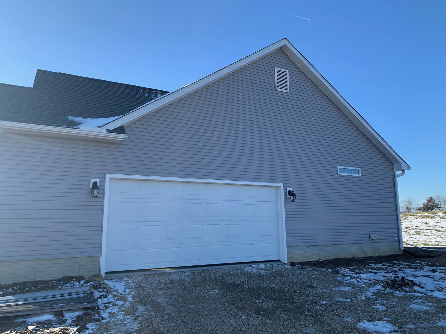 White garage door and gray roof on a house, concrete driveway in foreground, snow-covered yard and trees in background