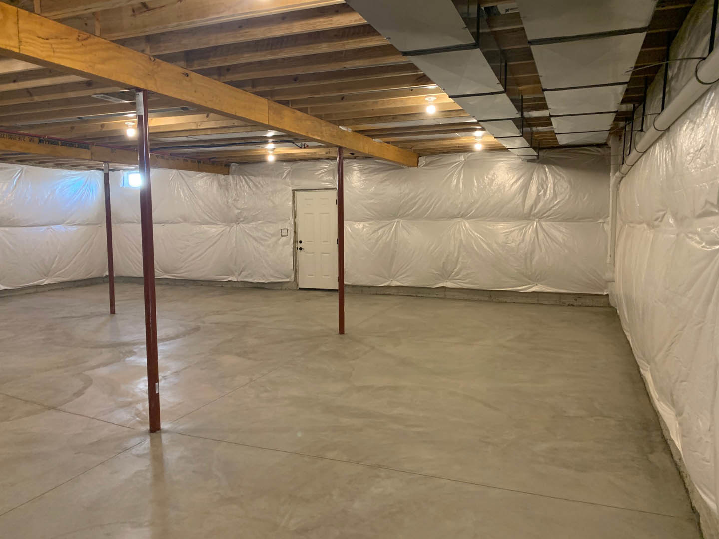 Room under construction with white plastic coverings draped over walls, concrete floor with exposed metal pole, white door featuring black knobs, ceiling with glass panel and