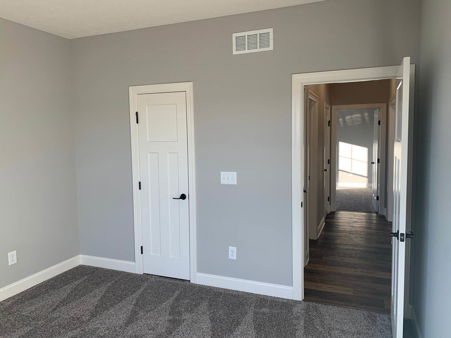 White walls with two paneled doors featuring black handles, dark wood laminate flooring illuminated by natural light, ceiling vent, white wall outlet, and a close-up of beige
