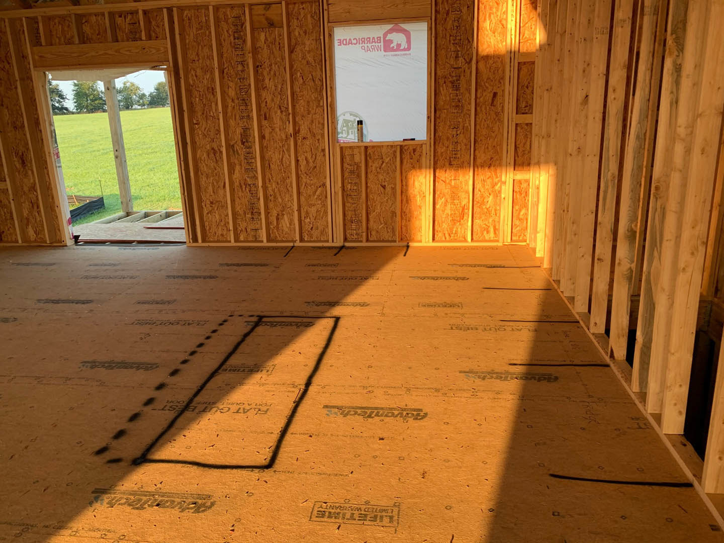 Sunlit room featuring wood plank flooring, white walls, large window with view of grass and trees, and a door; shadow cast across floor, white paper with red logo resting nearby.