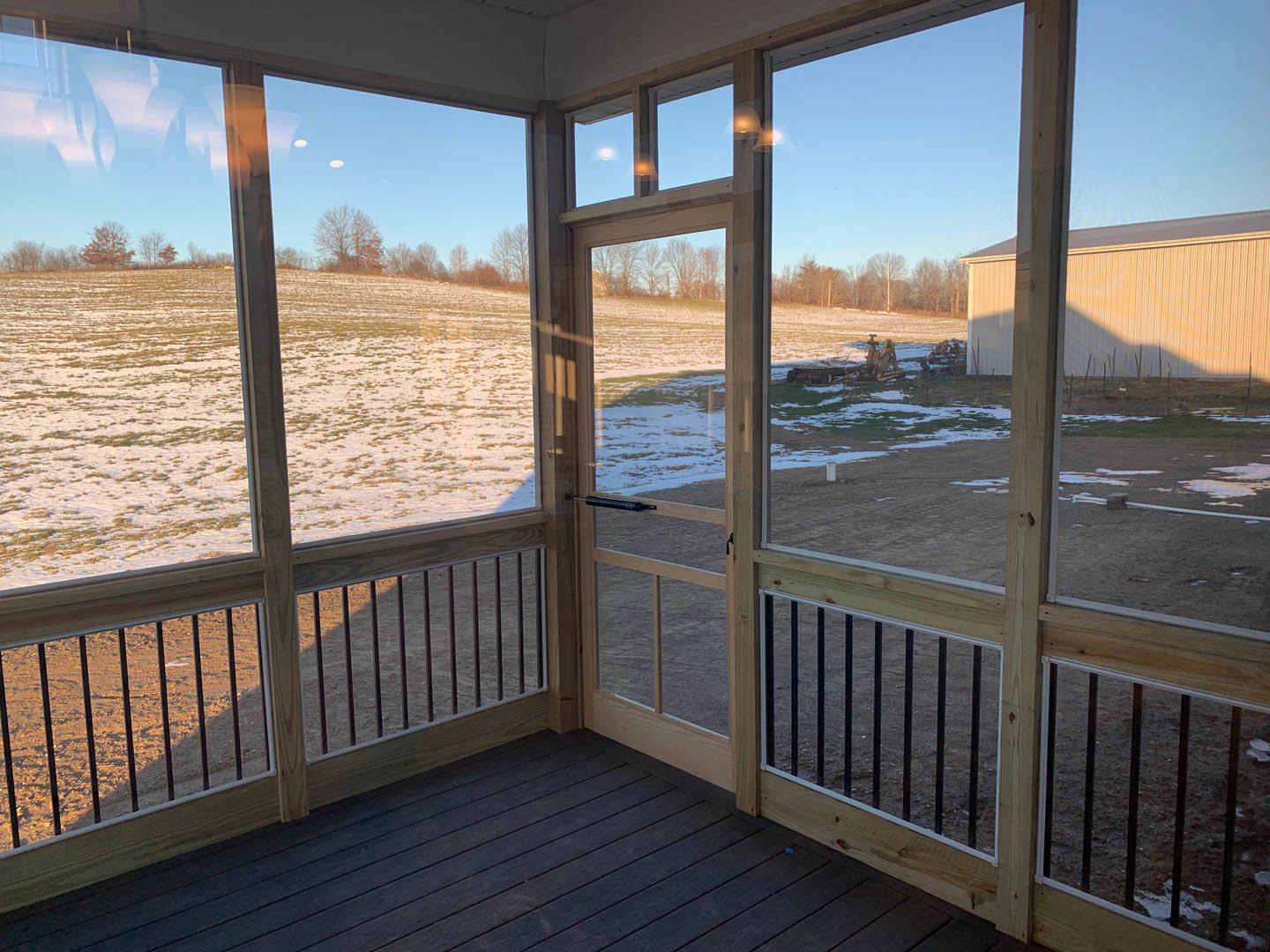 Screen door opening onto a snowy field, wooden porch floor in foreground, leafless tree visible through window against blue winter sky