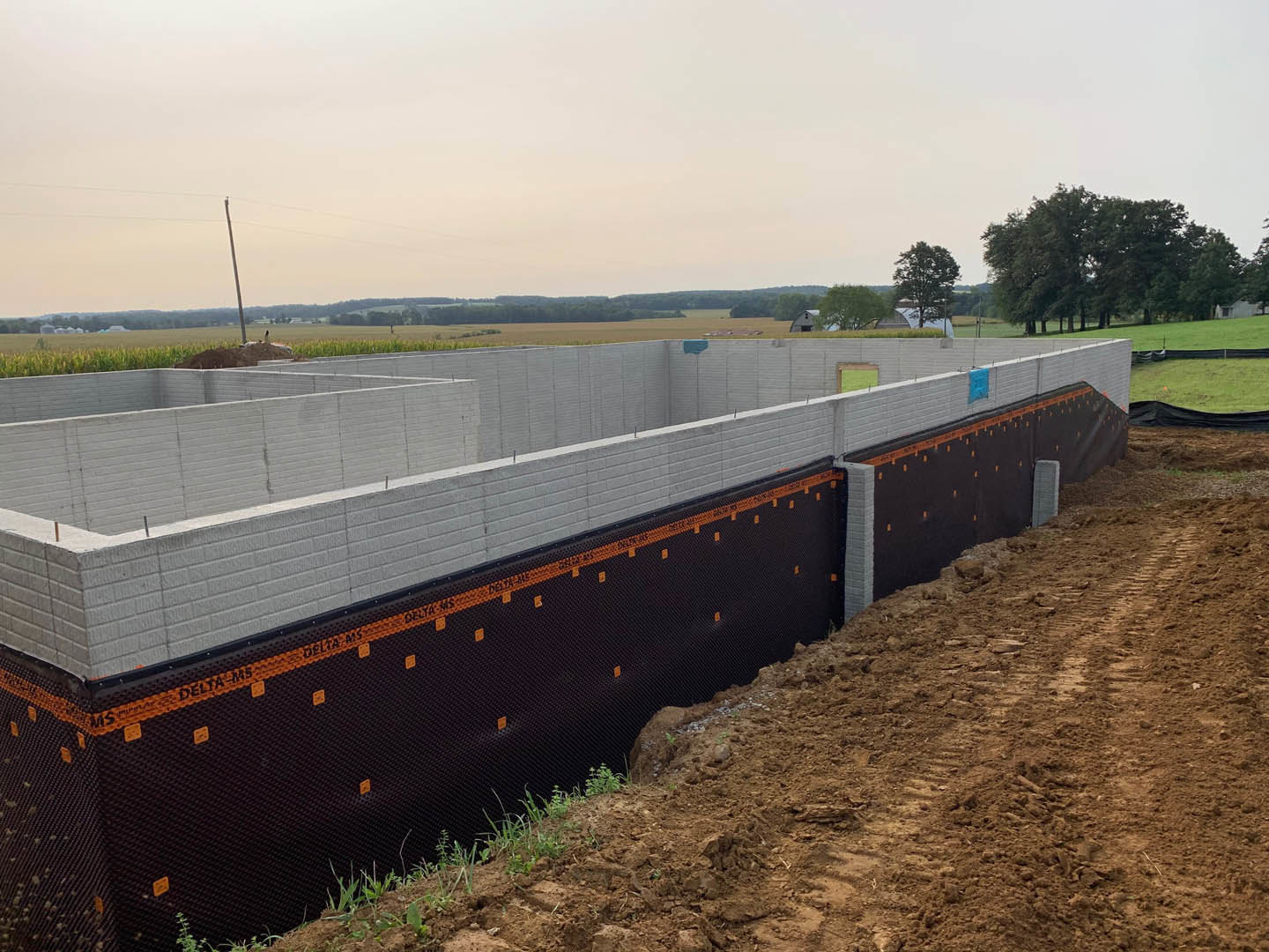 Black and orange construction wall beside white wall, dirt road bordered by black fence, grassy area with scattered trees under blue sky