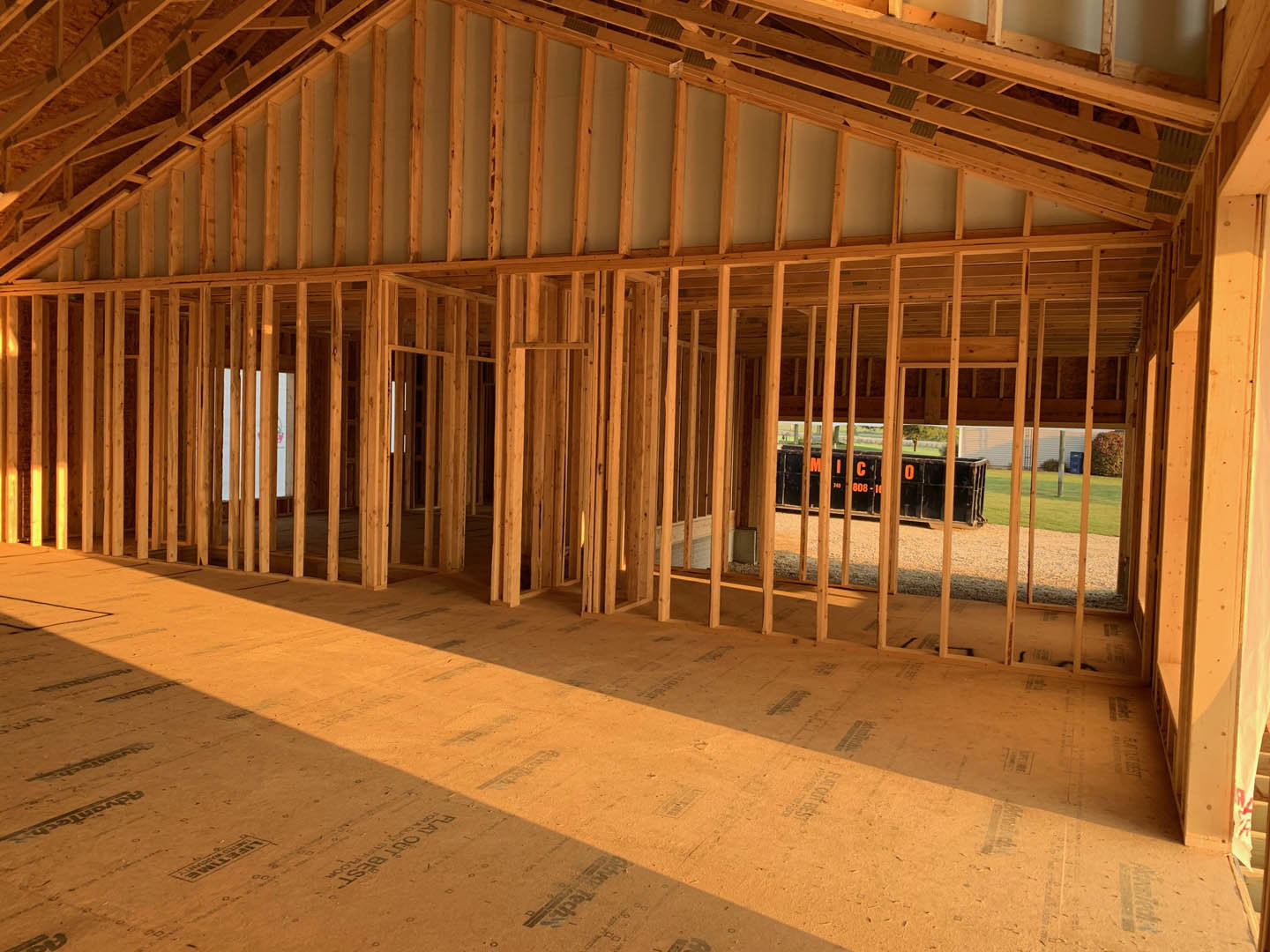 Wood framing and exposed beams inside a house under construction, open view toward grassy field, unfinished floors and walls with construction materials visible