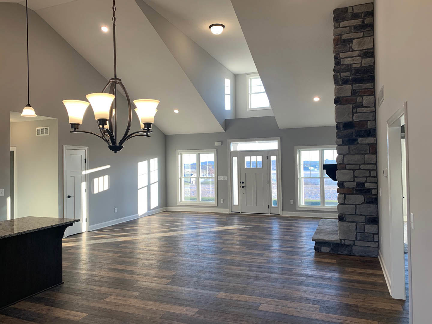Spacious living area with wood flooring, stone fireplace, chandelier, stone pillar beside window, and kitchen island; window overlooks open field.
