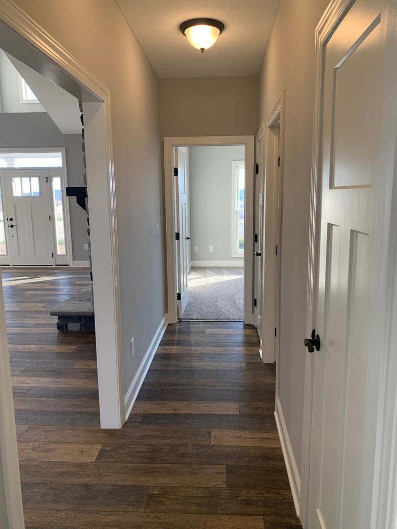 Hallway with white paneled doors, hardwood flooring, white baseboards, ceiling light fixture, open door revealing adjacent room, and partial view of a wooden table