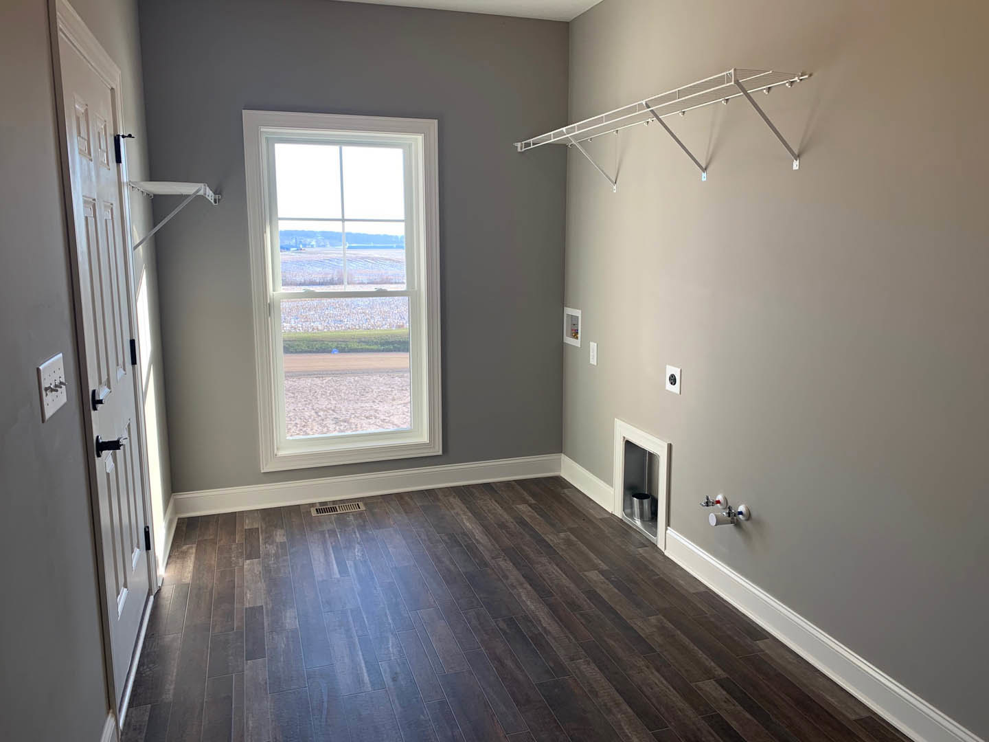 Sunlit room with wide window overlooking grassy field, wood plank flooring, white plaster walls, silver trash can near white door, metal bar mounted on wall, close-up of light